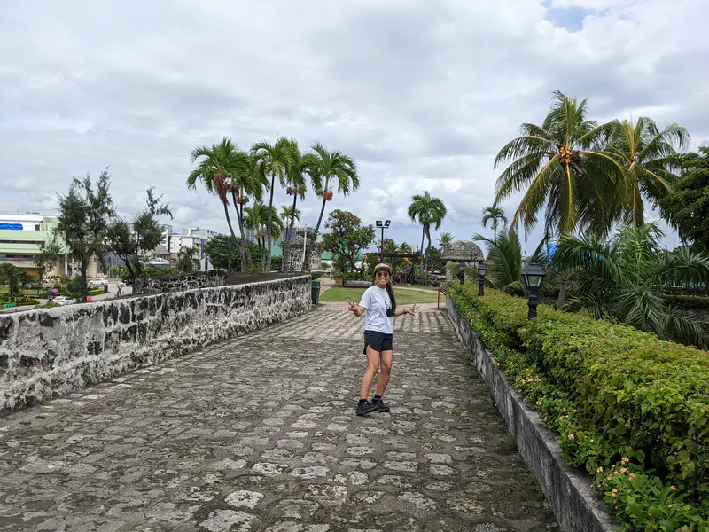 Person in a hat and white shirt standing on a stone pathway lined with palm trees and greenery.