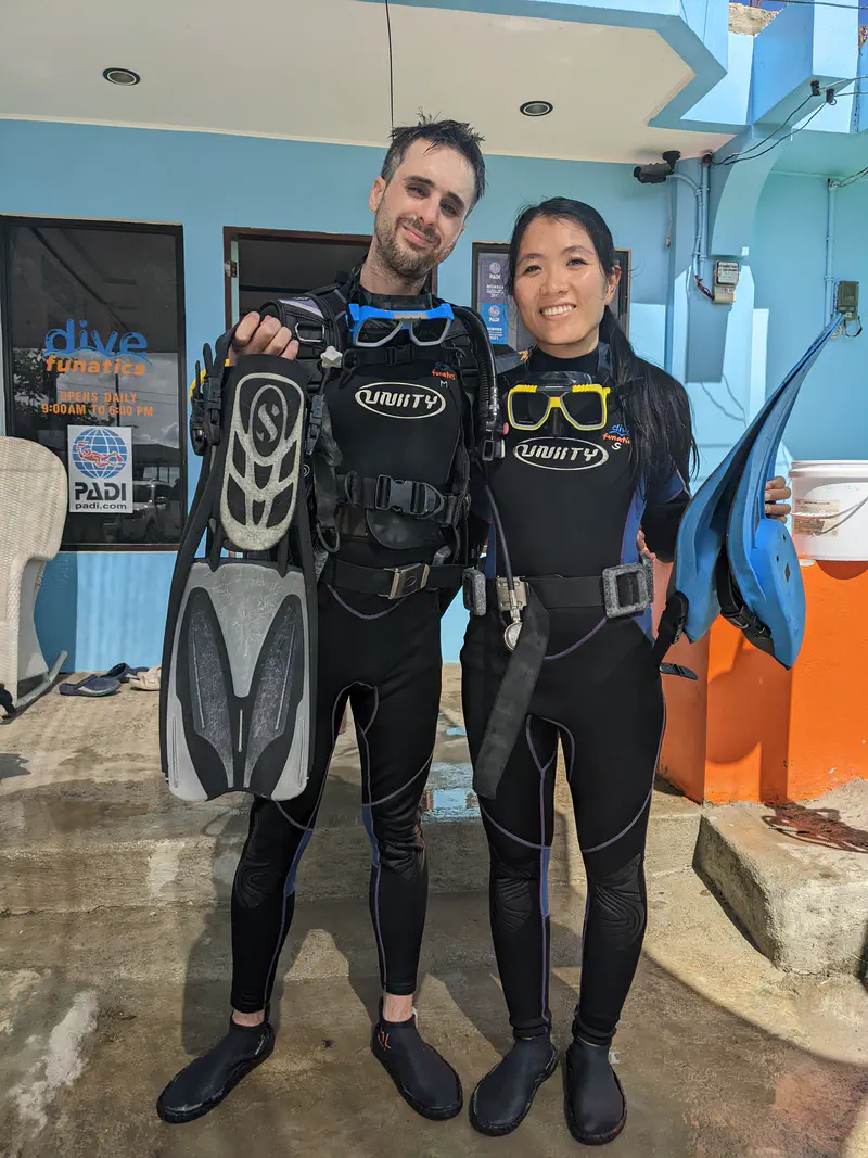 Two scuba divers in wetsuits and gear holding fins and masks, standing outside a dive shop.