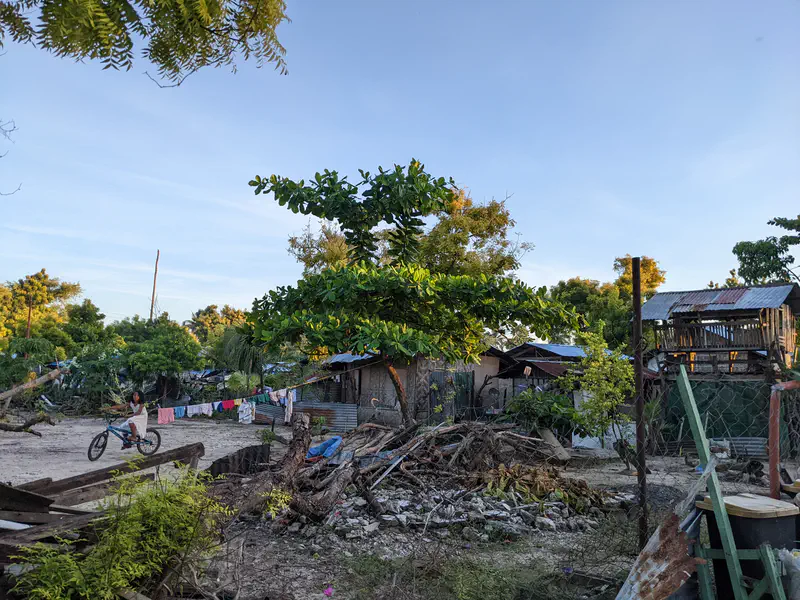 Child riding a bicycle near simple houses, laundry on a line, and scattered debris in a rural yard.
