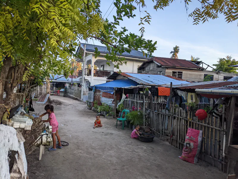 Children playing along a dirt path lined with simple houses and laundry hanging outside.