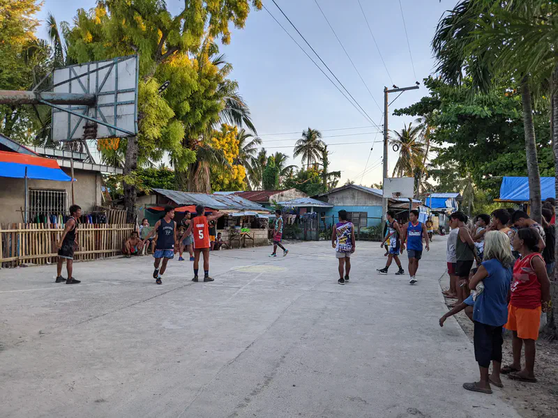 Group of men playing basketball on a street court surrounded by spectators in a village setting.