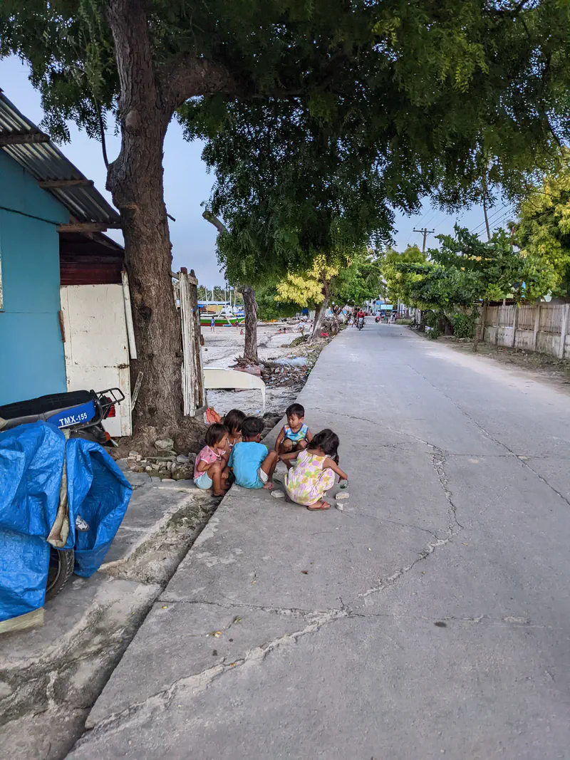 Group of children sitting on the roadside under a tree near houses and boats in the distance.