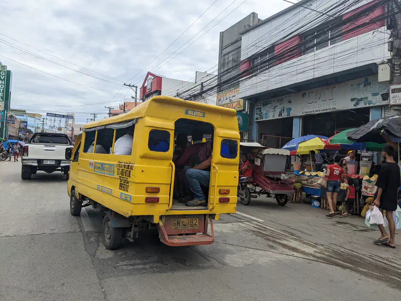 Yellow jeepney driving on a busy street with market stalls and pedestrians on the side.
