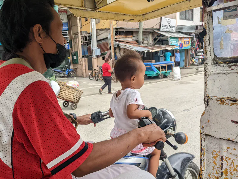 Man driving a motorcycle with a small child sitting in front, seen from inside a tricycle.