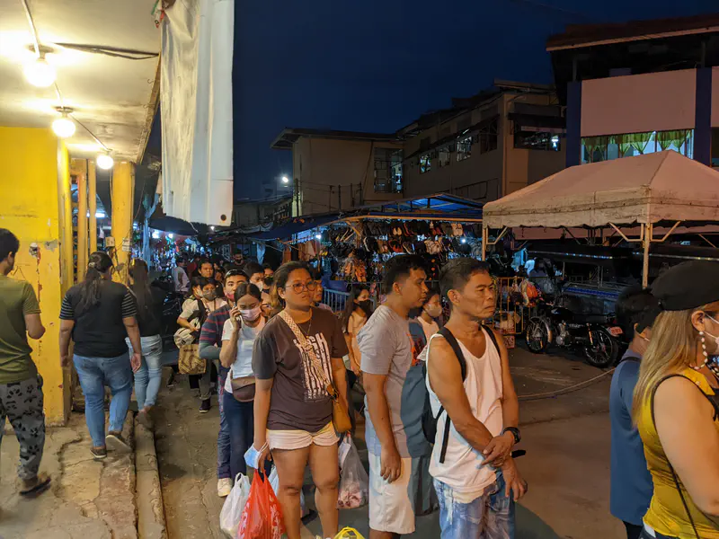 People lined up at night in a busy market area with stalls and bright lights.