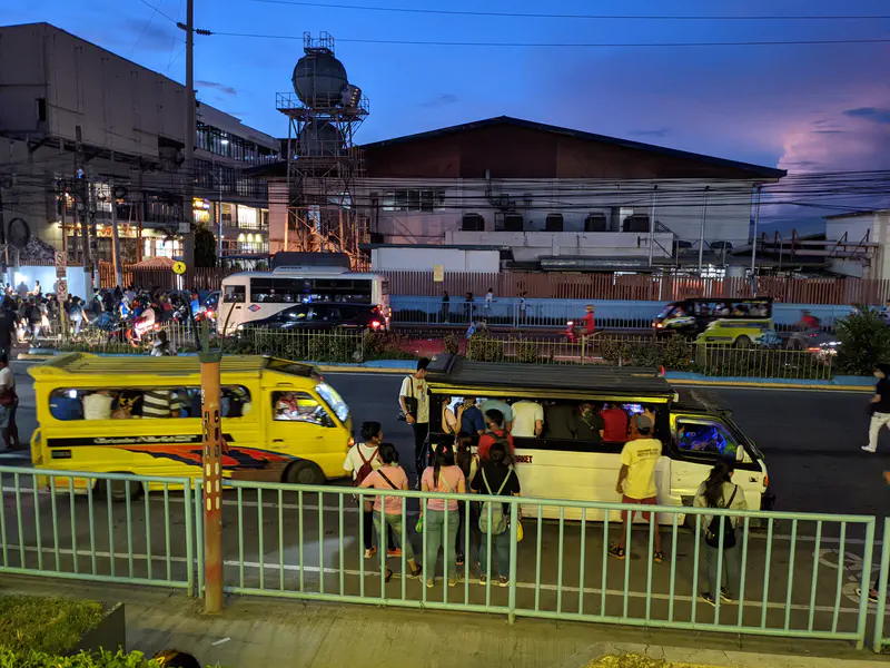 Commuters boarding jeepneys on a busy street at dusk, with traffic and buildings in the background.