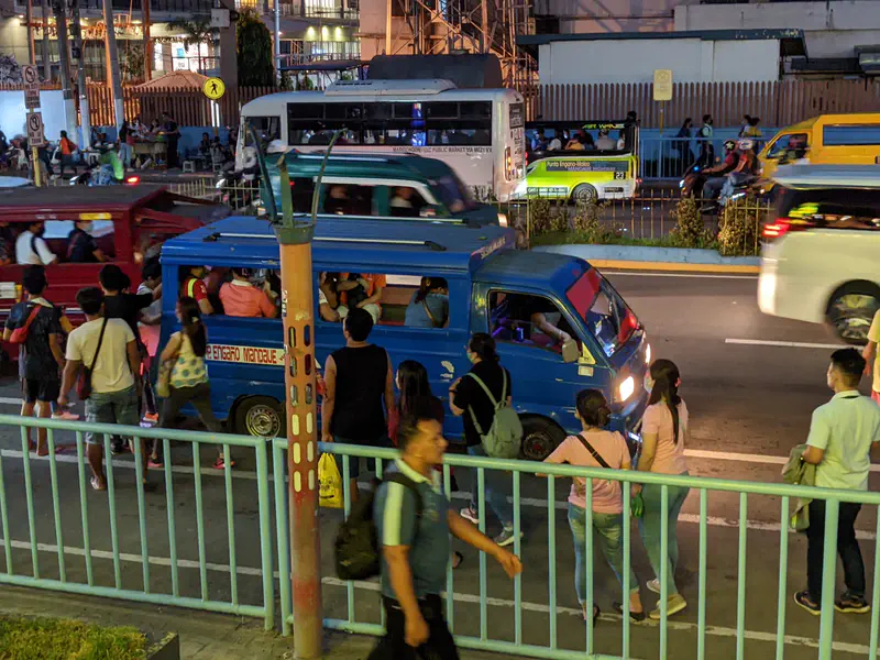 Commuters boarding a blue jeepney at a busy street in the evening, with other jeepneys and vehicles passing by.