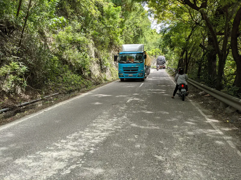 A blue cargo truck and a tanker driving on a narrow mountain road with trees and motorcycles nearby.