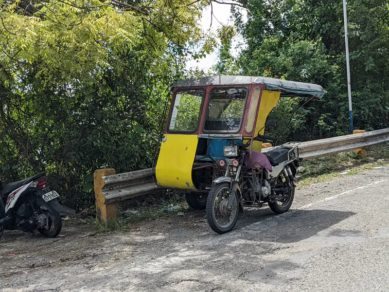 A tricycle with a yellow and blue sidecar parked by a guardrail under trees.