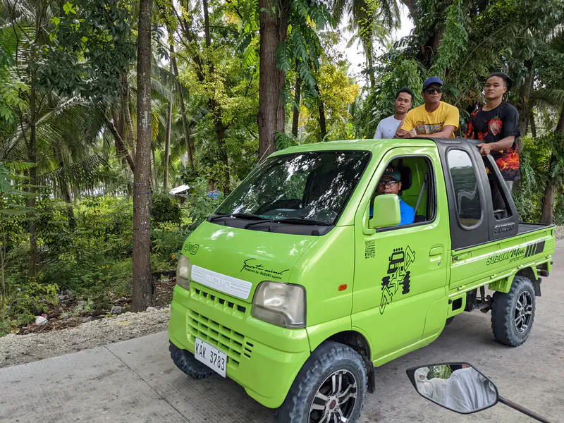 Group of young men riding in the back of a bright green mini truck on a rural road.