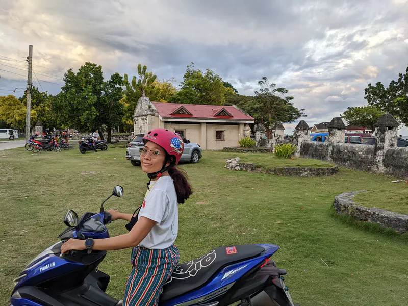 A person wearing a pink helmet sitting on a blue Yamaha scooter in a grassy area near old stone structures and trees.