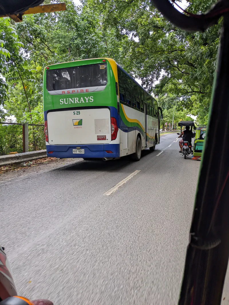 A green, white, and blue Sunrays bus driving on a tree-lined road with tricycles nearby.