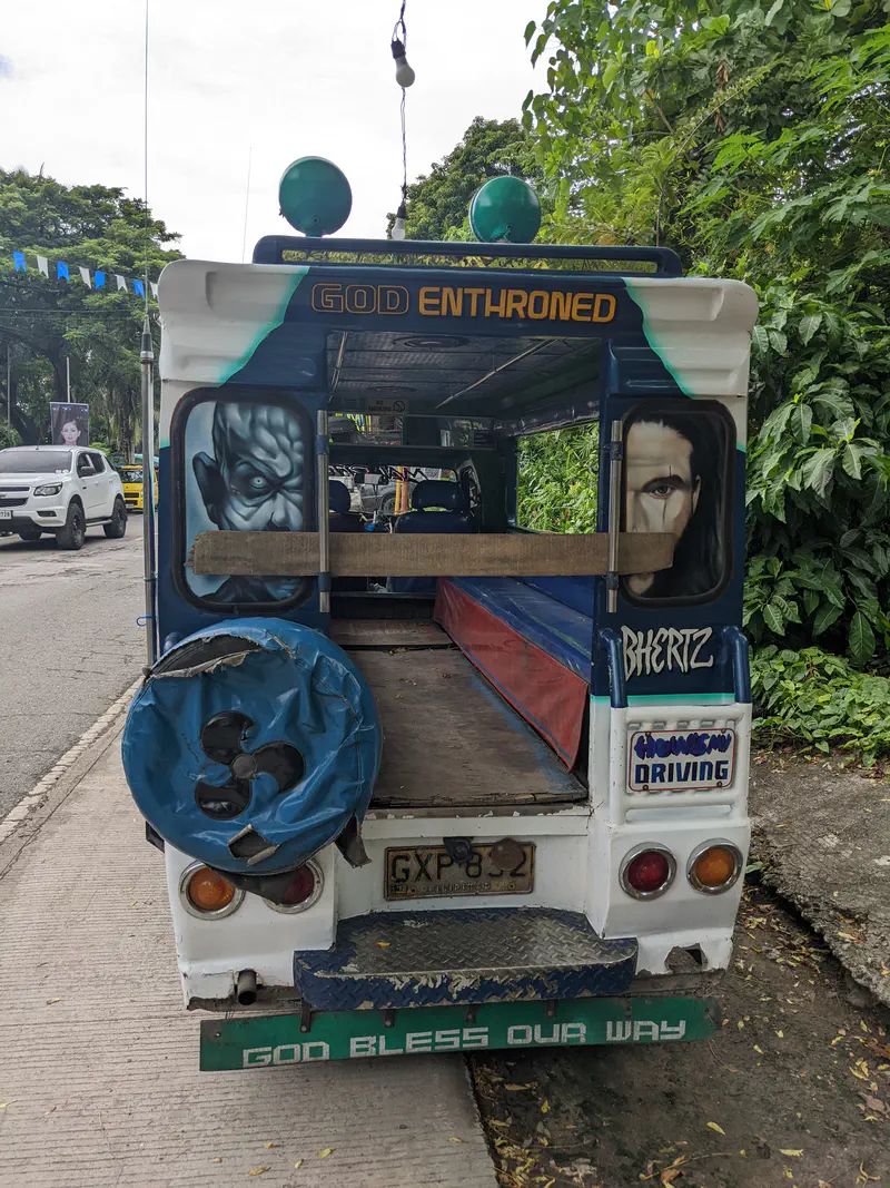 Rear view of a jeepney with religious artwork and text reading 'God Enthroned' and 'God Bless Our Way,' showing wooden benches inside.