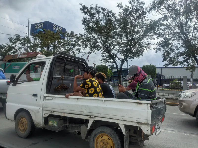 Several people sitting in the back of a small white pickup truck in traffic.