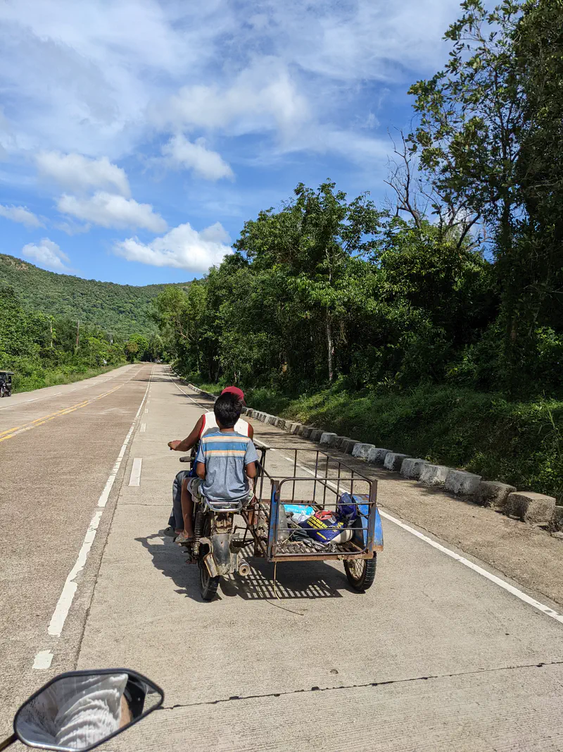 Two people riding a motorcycle with a metal sidecar carrying items along a rural road with trees and mountains.