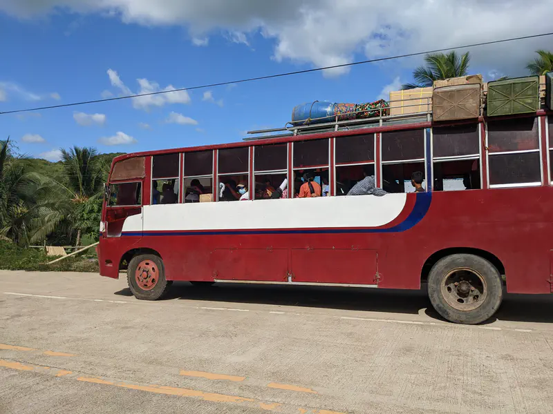 A red provincial bus with passengers inside and luggage tied on the roof, traveling on a sunny rural road.
