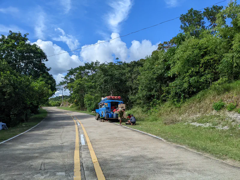 A blue jeepney stopped on the side of a rural road surrounded by greenery, with people gathered near it under a bright blue sky.