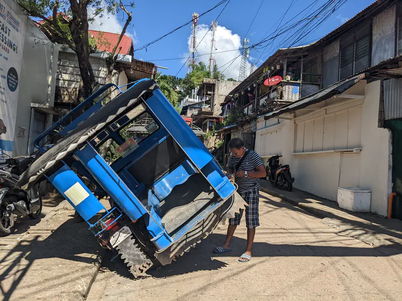 A man checking a blue tricycle tilted on its side in a narrow residential street on a sunny day.
