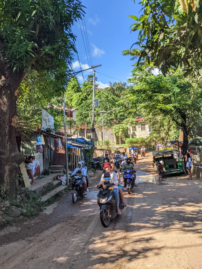Motorcycles and a tricycle driving along a narrow, muddy road in a residential area with trees and houses.