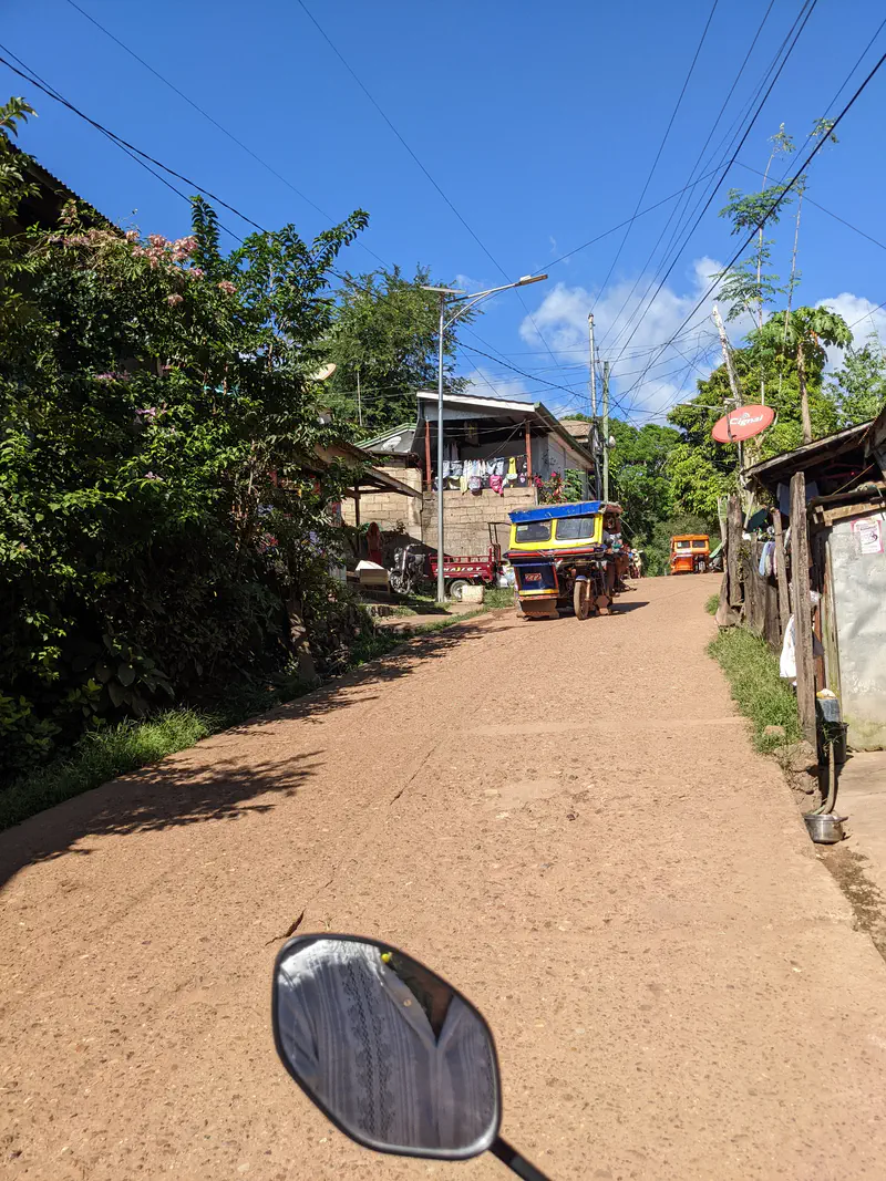 A yellow tricycle driving uphill on a narrow street in a residential area with houses and laundry hanging outside.