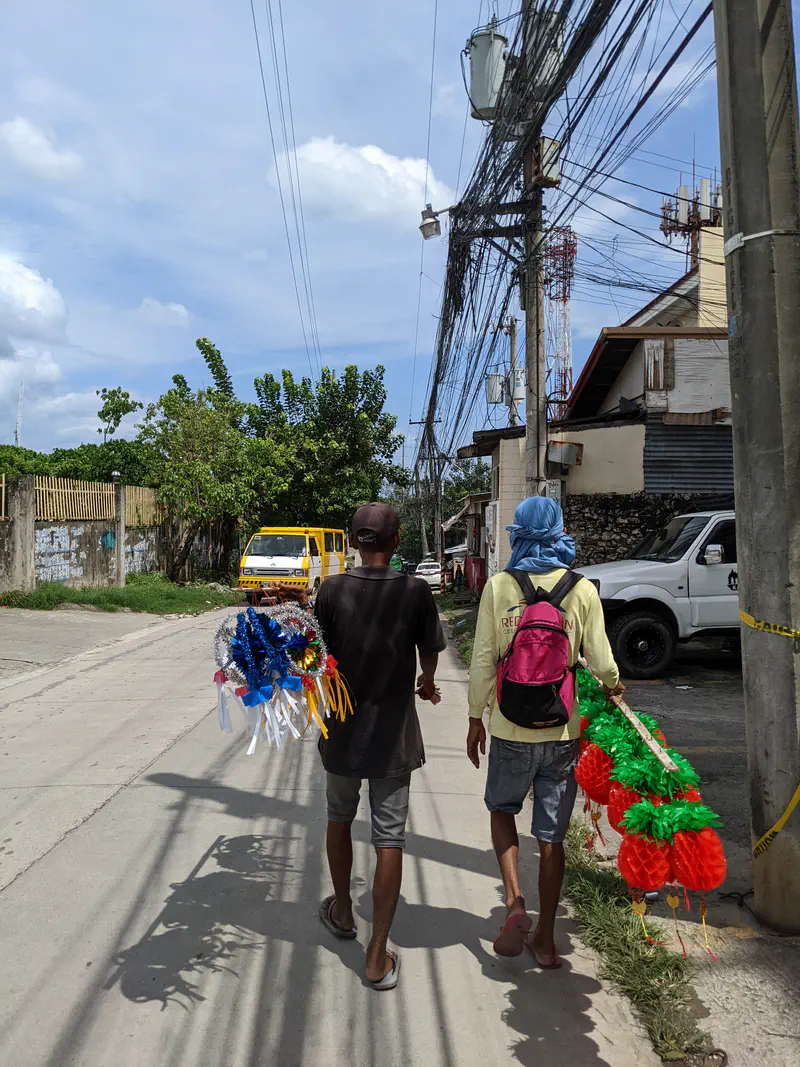 Two men walking on a street carrying colorful decorations under tangled electric wires.