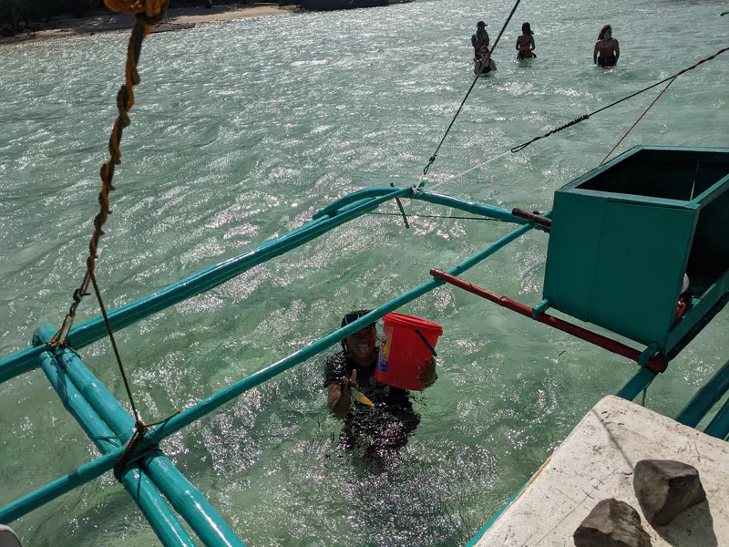 Person standing in the sea holding a red bucket near a boat, with people in the water and beach in the background.