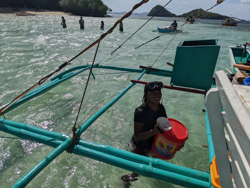 Woman standing in shallow sea water holding a red bucket beside a boat, with small islands and people swimming in the background.