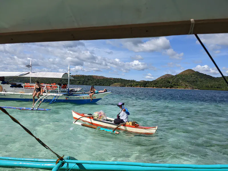 Person in a small wooden boat selling goods near a larger tour boat with tourists by the shore of a tropical island.