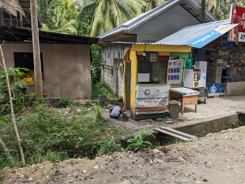 Tiny roadside store with lottery signage beside a child playing on the ground.