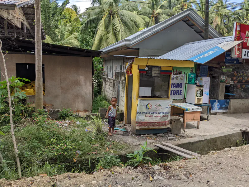 Young child standing barefoot near a small roadside store with lottery signage.