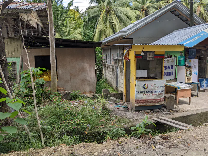 Yellow roadside lottery store beside older buildings and lush greenery.