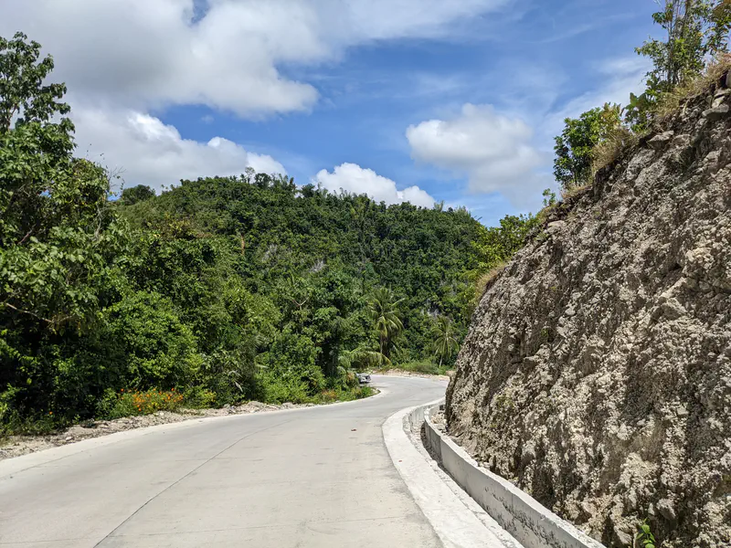 Curved concrete road passing through green hills and rocky cliffs.