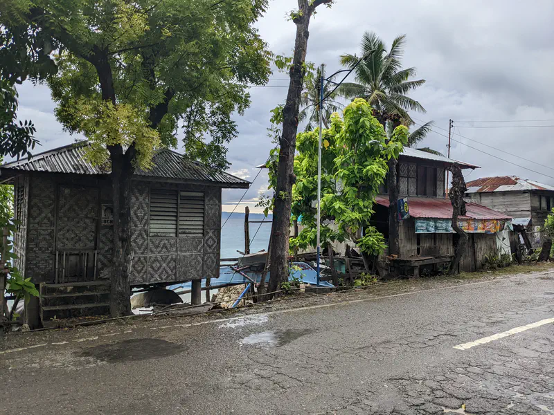Wooden houses on stilts by the sea beside a wet roadside with palm trees.