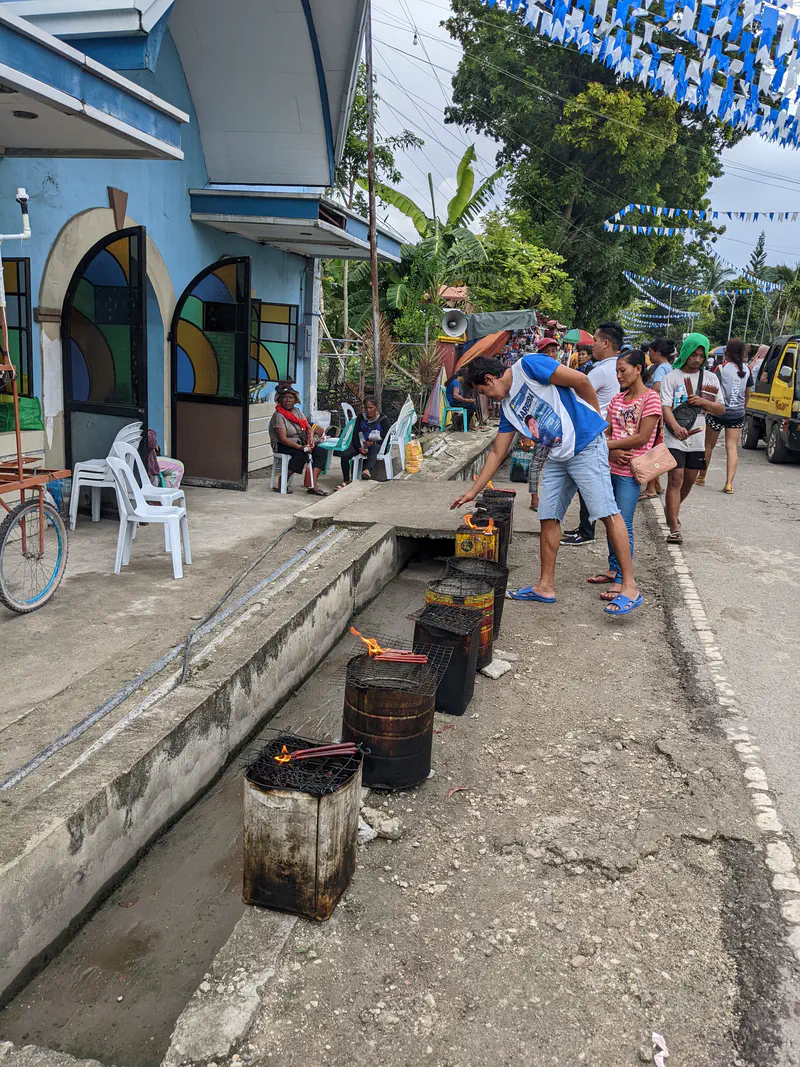 People lighting candles on metal barrels outside a church during a fiesta celebration.