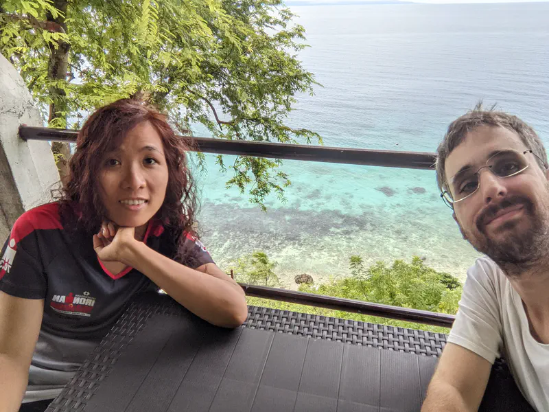 Two people taking a selfie at a seaside table overlooking clear blue water.