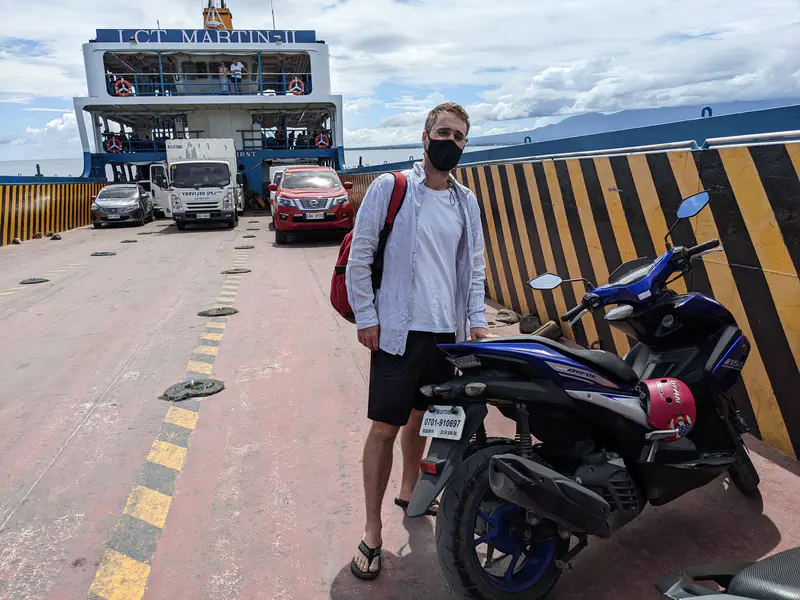 Person with a motorbike standing on a ferry deck with cars and trucks in the background.