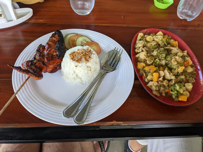 Plate with grilled chicken, rice, and cucumber slices beside a dish of mixed vegetables.