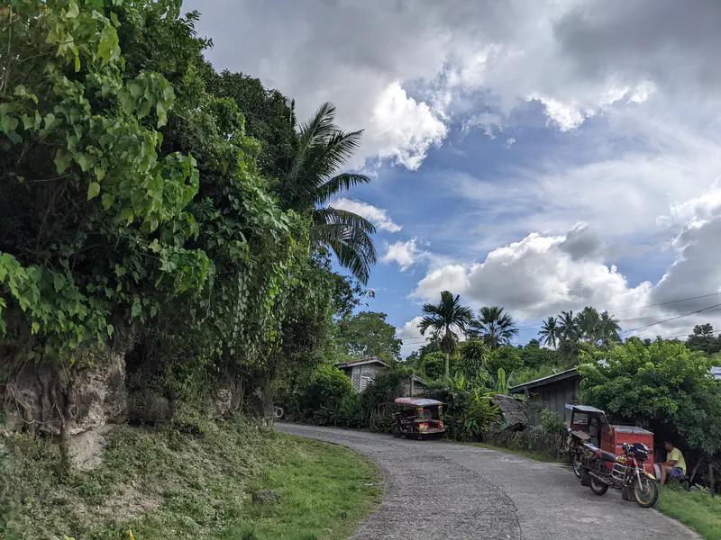 Narrow road curving through a village with tricycles parked beside houses and lush greenery.