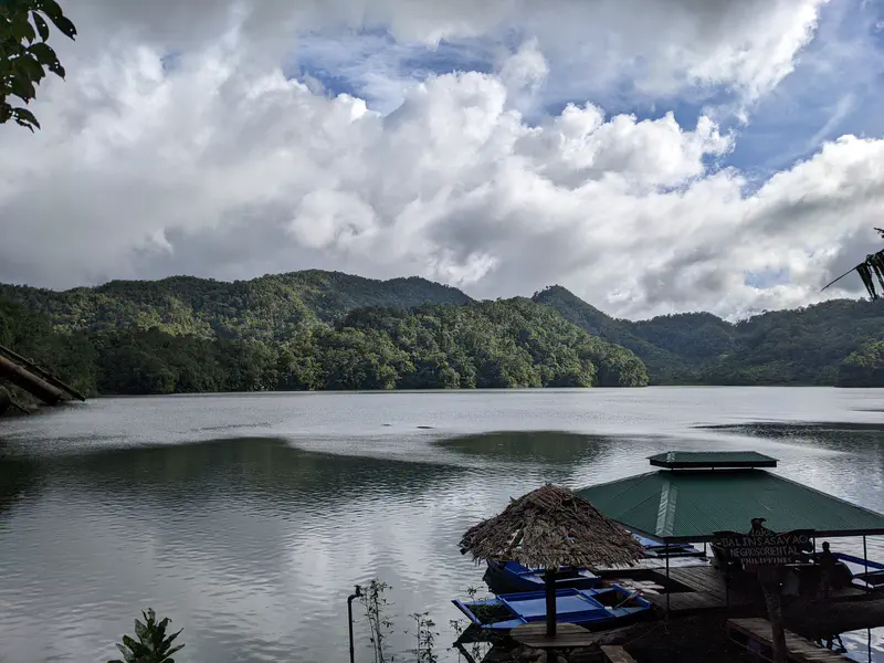 Mountain lake with dense green hills under cloudy skies and a floating hut by the shore.