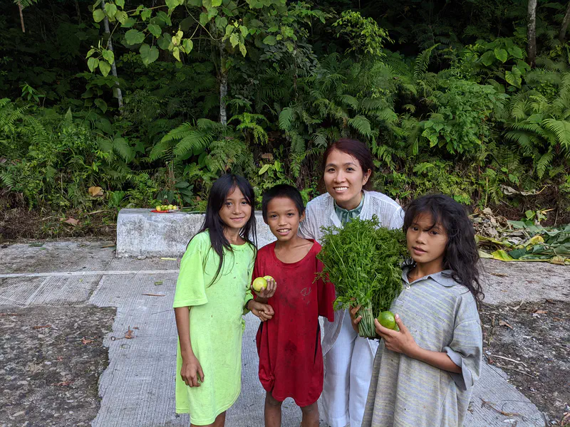 Woman smiling with three children holding vegetables and fruits by a roadside.