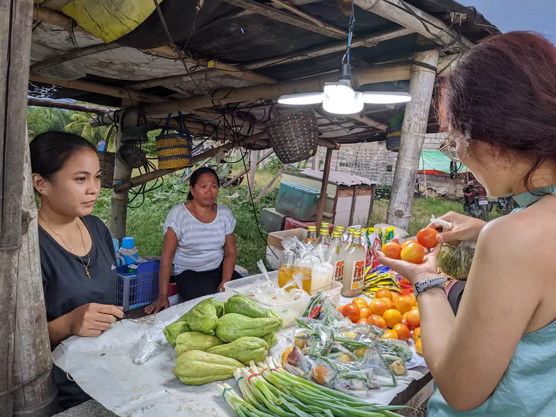 Woman buying tomatoes at a small market stall with vegetables and bottled goods.