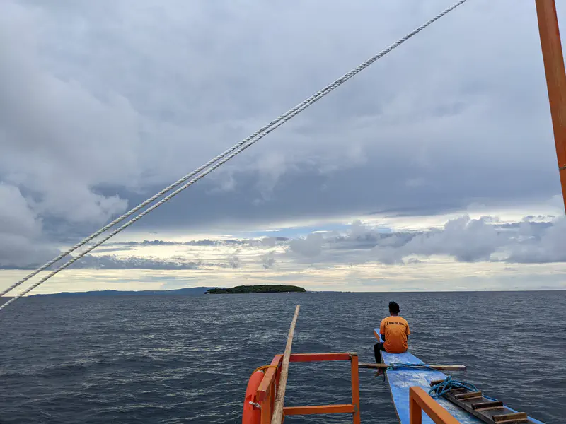 Person in an orange shirt sitting at the edge of a boat looking toward a distant island.