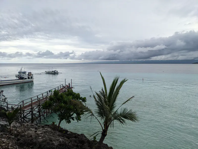 Wooden pier stretching into calm turquoise water with boats anchored nearby under cloudy skies.