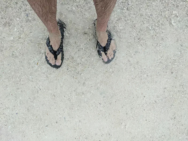 Feet in black flip-flops standing in shallow clear water on sandy ground.