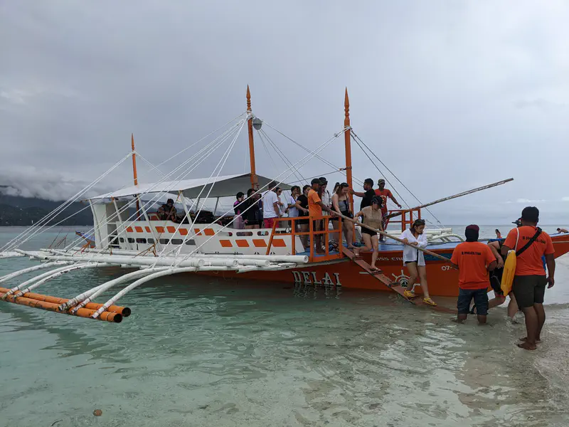 Group of people boarding an orange outrigger boat from shallow water.