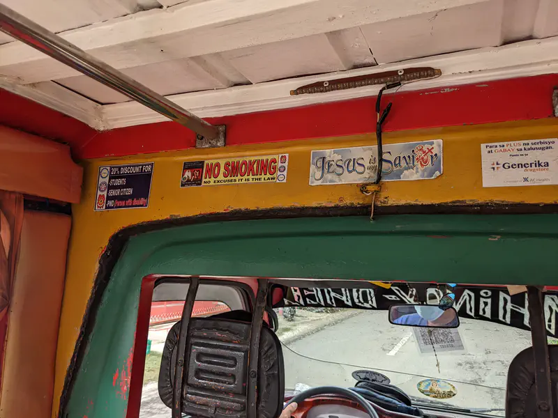 Inside a jeepney with signs including 'No Smoking' and 'Jesus is Savior' above the driver's seat.
