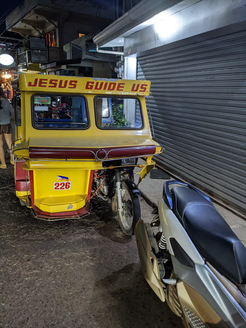 Yellow tricycle with a sign reading 'Jesus Guide Us' parked on a street at night.