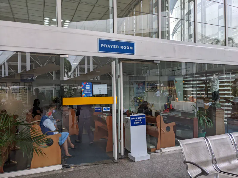 Glass-walled prayer room with pews, statues, and people seated inside.