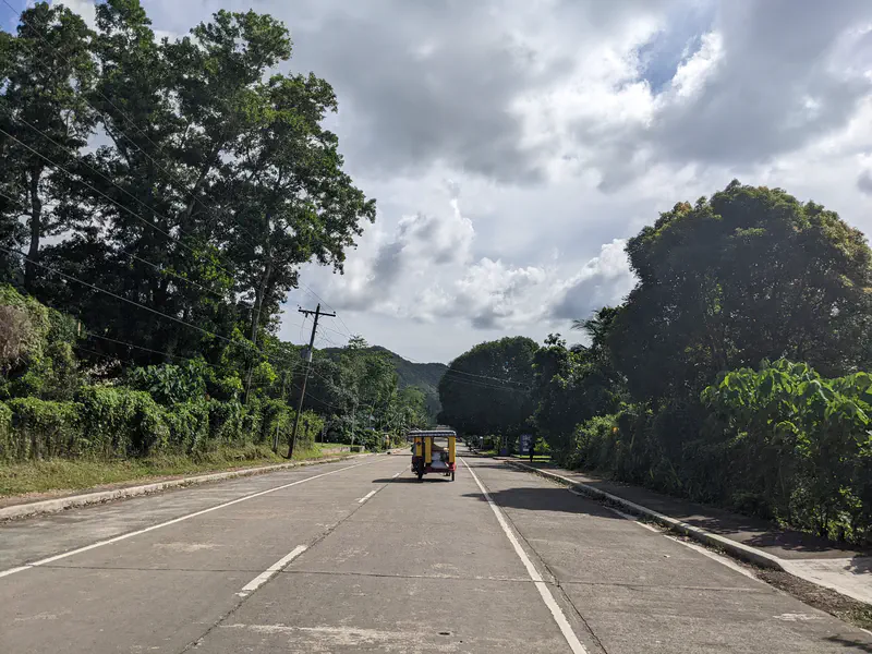 Yellow tricycle driving along a wide road lined with trees under a cloudy sky.