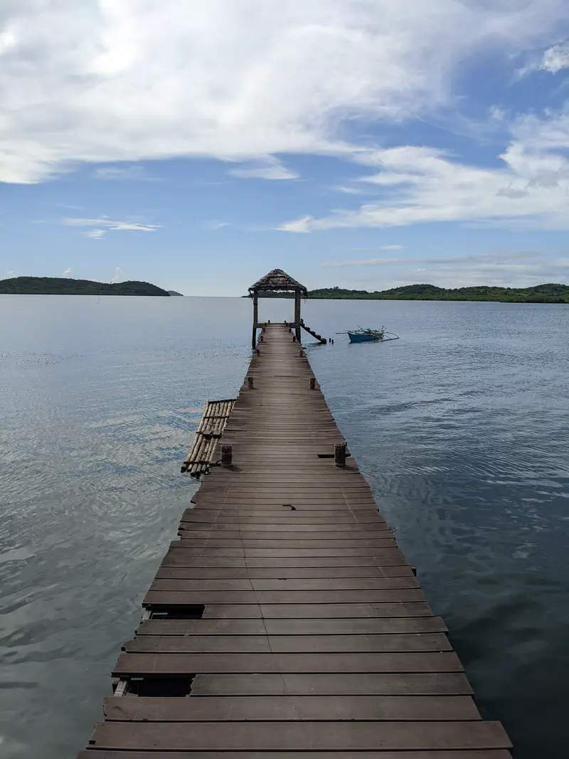 Wooden pier with a small shelter extending into calm sea under a partly cloudy sky.